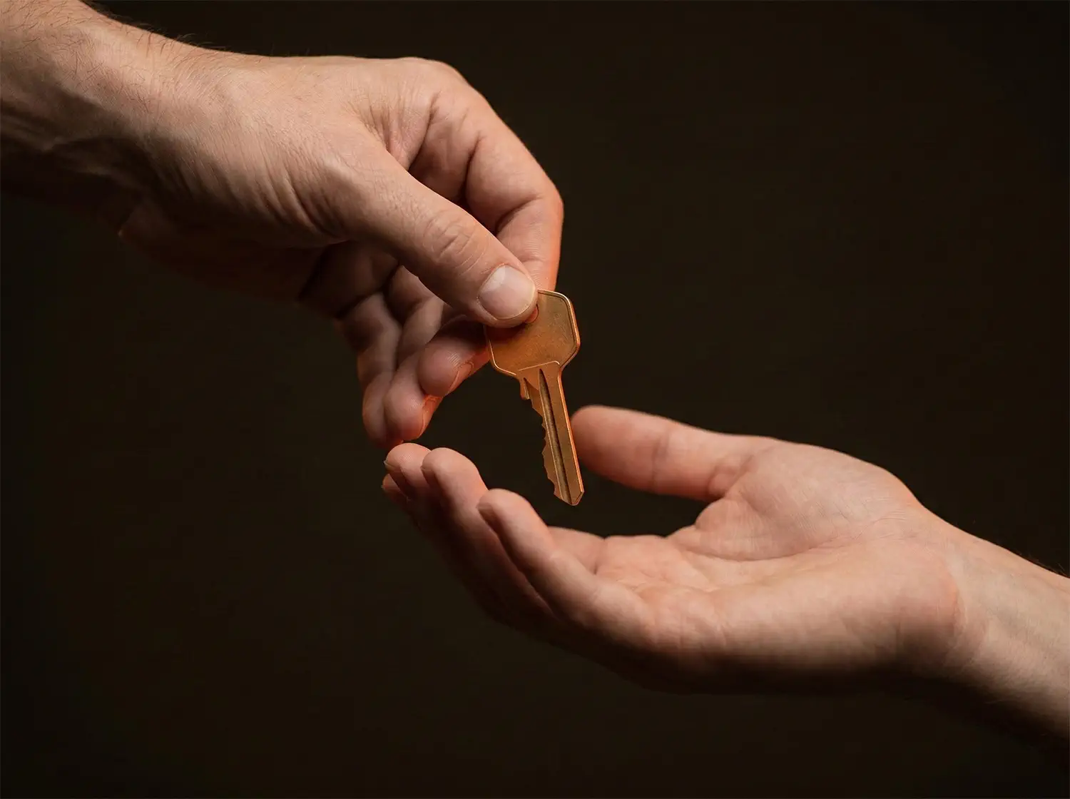 Close-up of two hands exchanging a warm brass key against a dark background