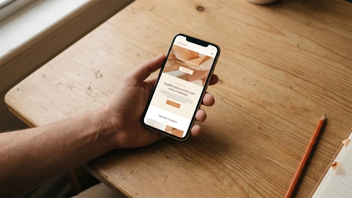 Hand holding a smartphone above a clean oak desk with warm-toned screen and burnt-orange pencil